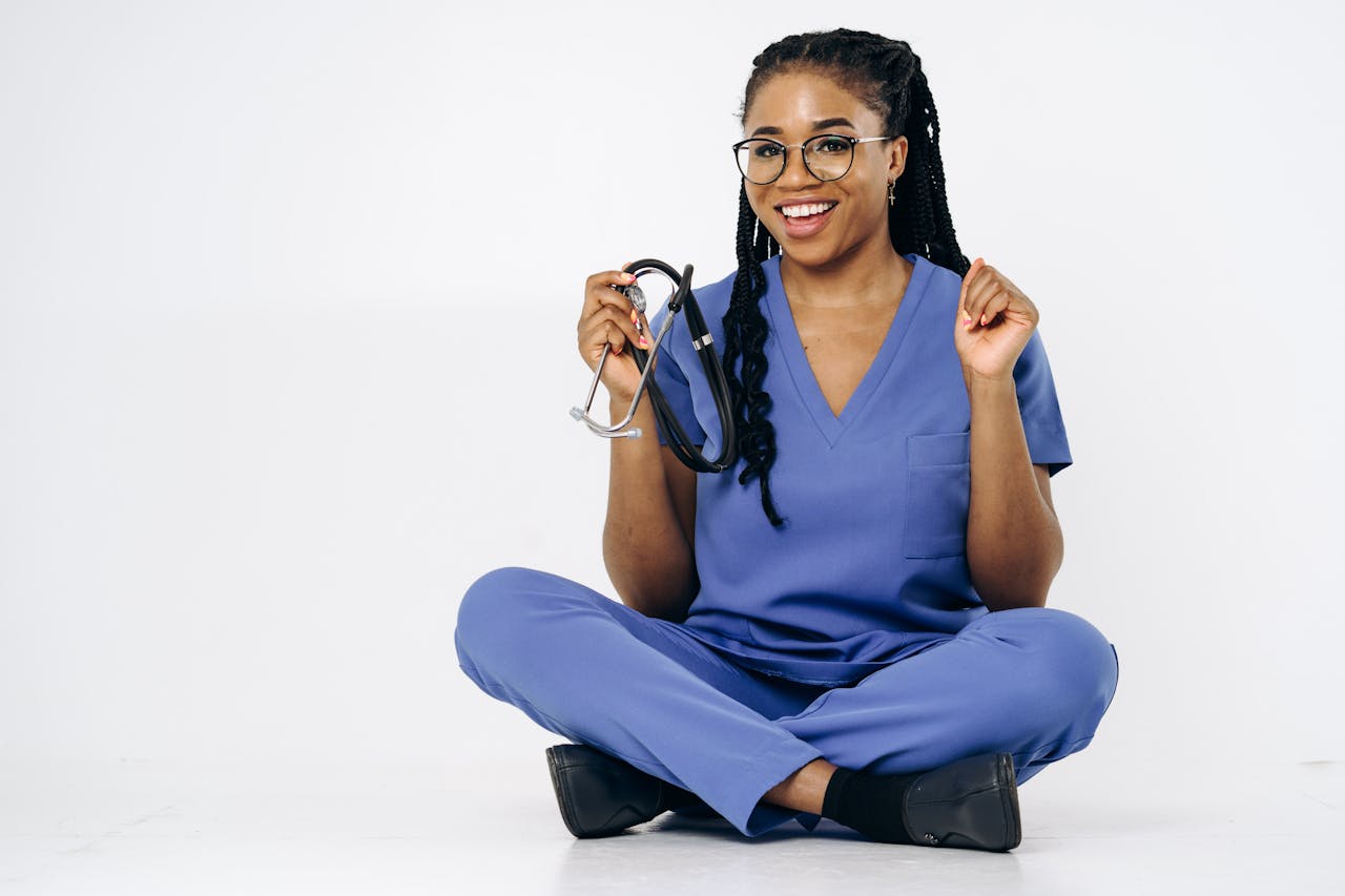 A cheerful nurse in blue scrubs holding a stethoscope while sitting, isolated on white.